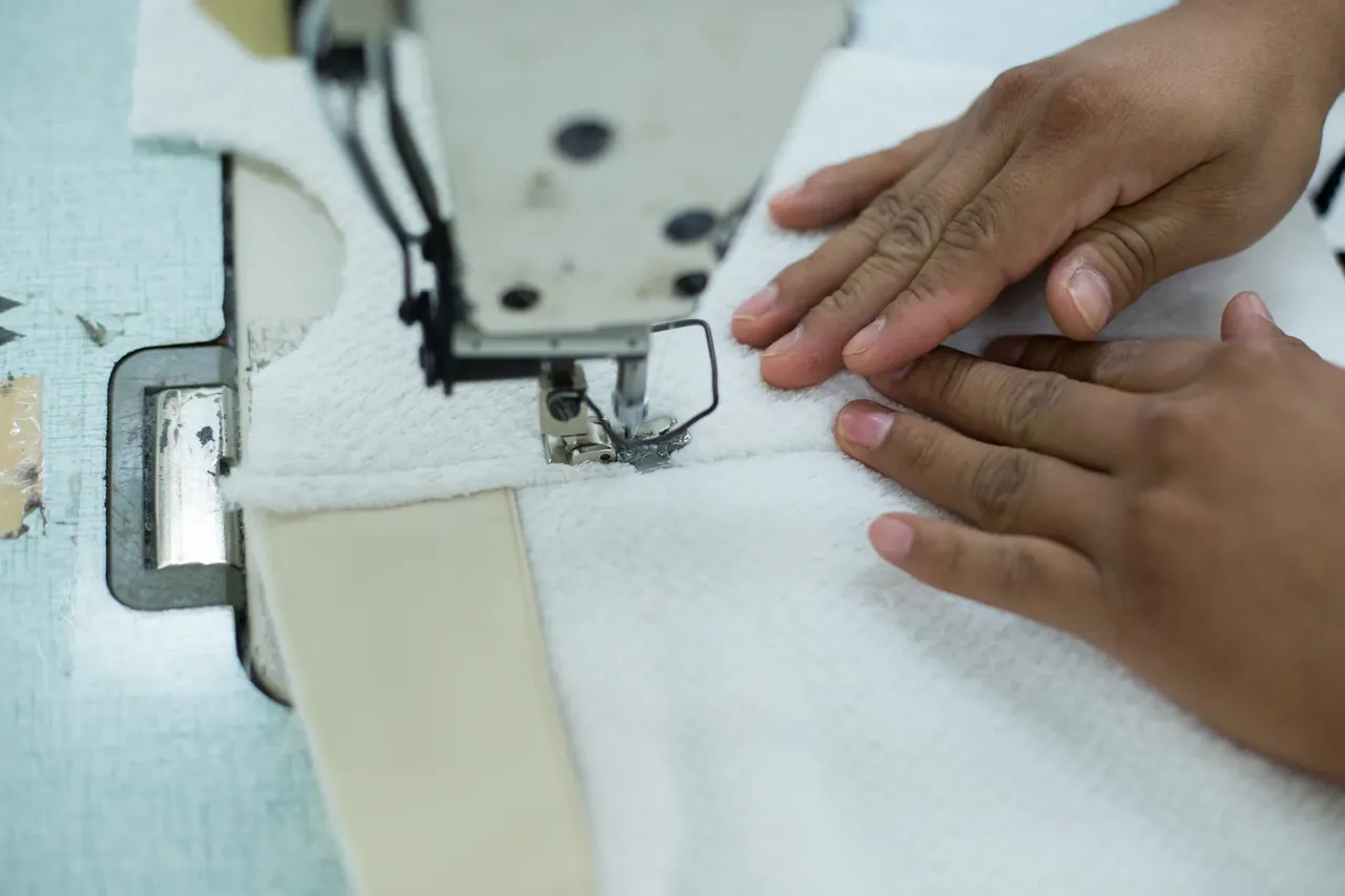 A machinist's hands guiding textured white fabric and a cream binding tape under the presser foot of an industrial sewing machine.