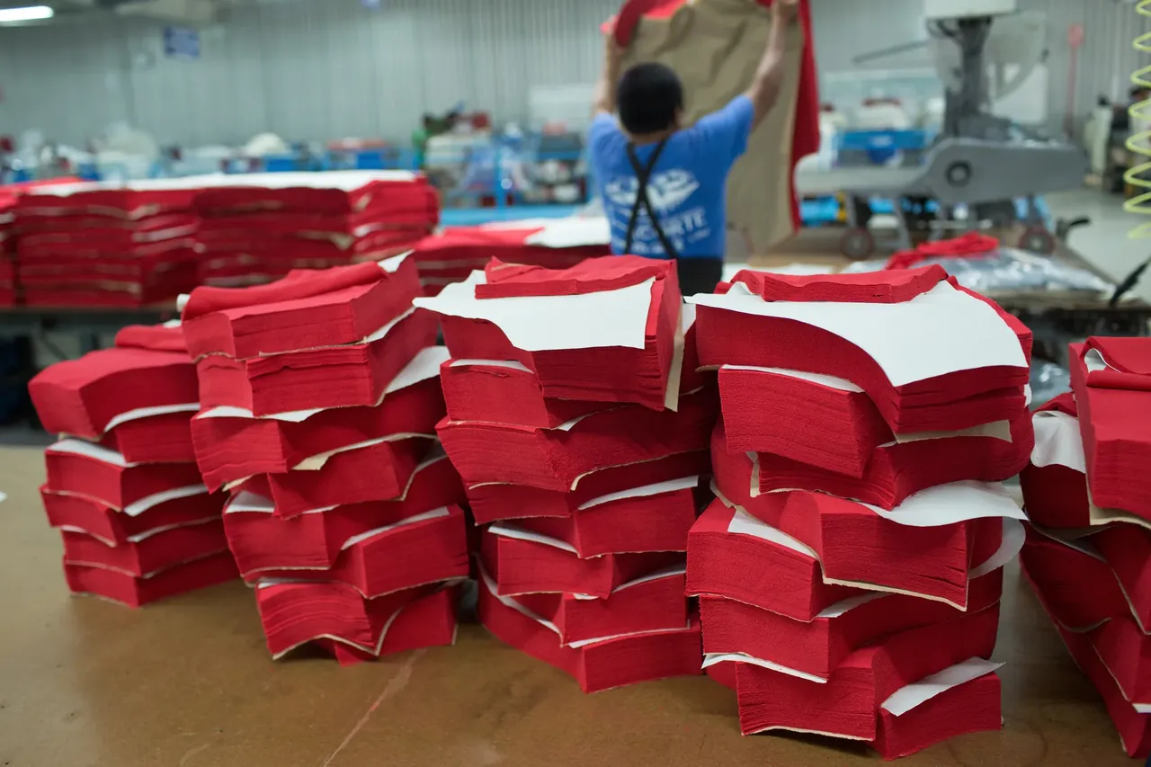 Stacks of cut red and white fabric pieces on a cutting-room table, with a worker bundling more at a bench behind.